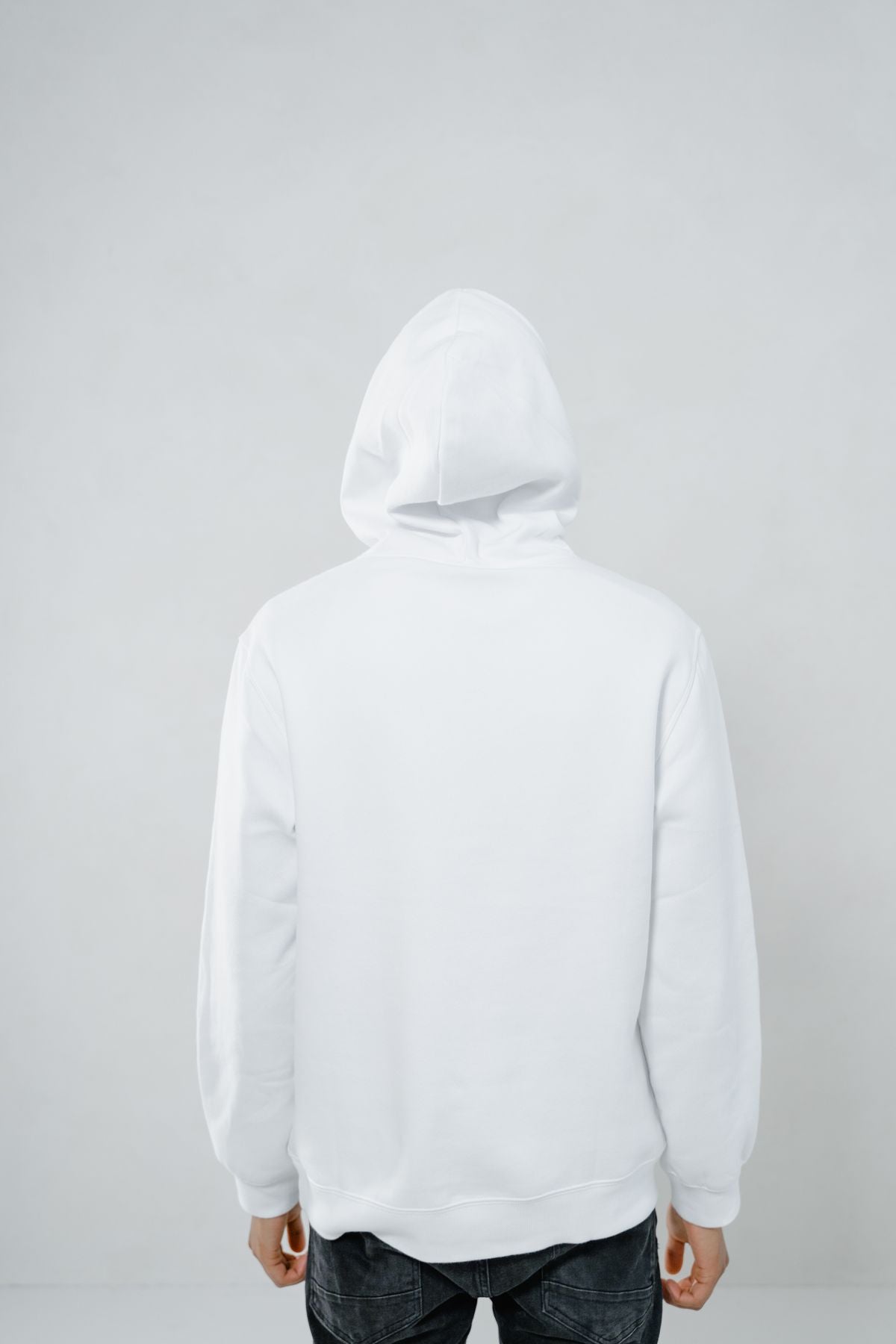 Back view of a person wearing a plain white pullover hoodie with the hood up, standing against a plain light grey background. Face not visible.